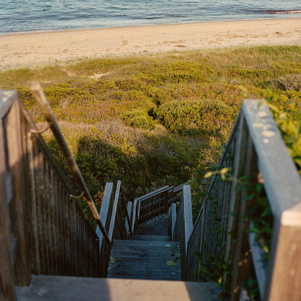 Weathered wooden stairway with handrails leads through dunes to a serene beach, embodying classic, effortless coastal charm and inviting a high-end, fun escape.