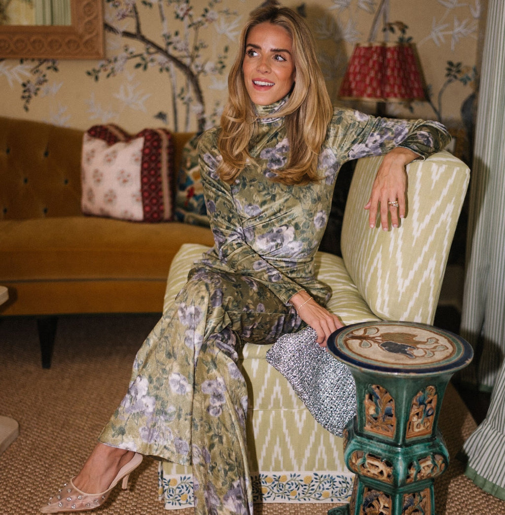 Woman in a classic green floral outfit with studded heels, holding a sparkly clutch, seated in an effortless, high-end, richly decorated living room.