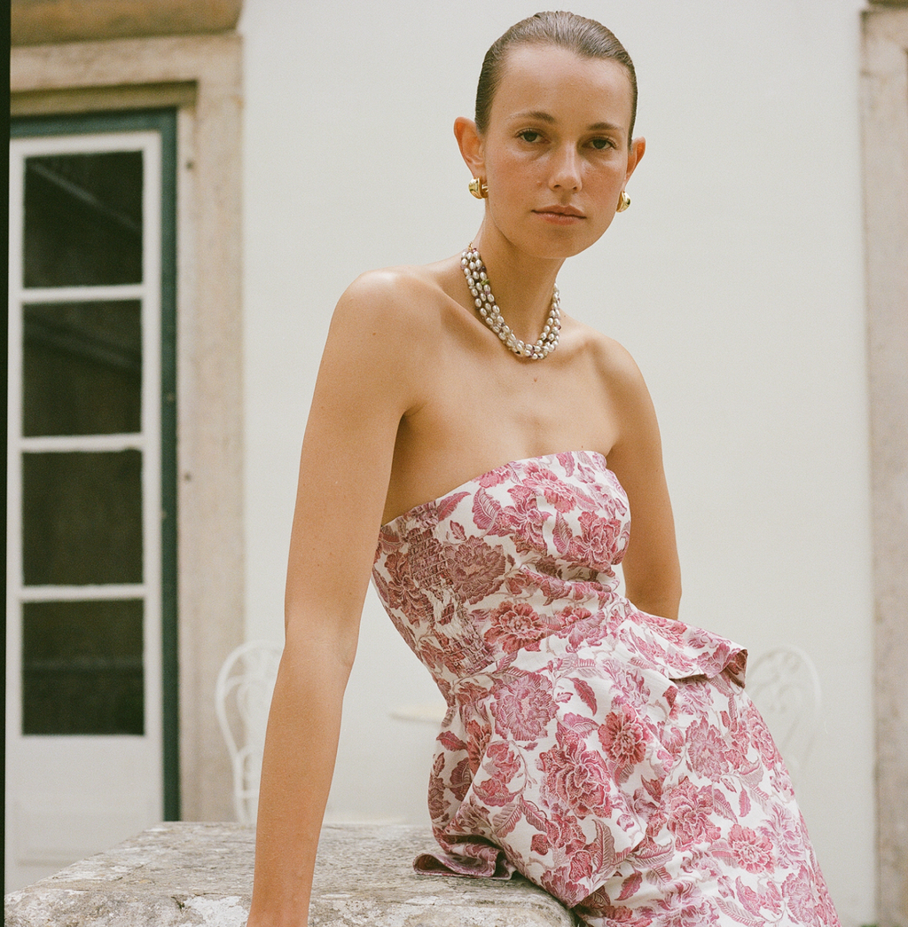 Young woman in a classic, strapless floral dress with pearl necklace and gold hoops, embodying effortless, high-end style on a sunlit terrace.