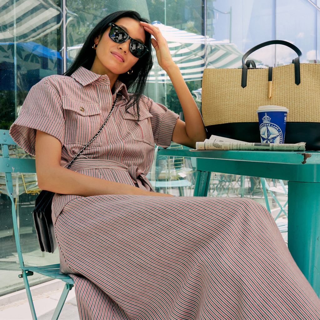 Young woman in sunglasses, classic pink-striped shirt-dress, seated at outdoor café. Straw tote, coffee, and newspaper on teal table, embodying effortless, high-end style.