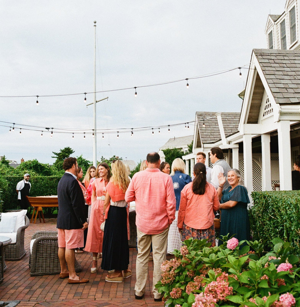 Casual outdoor gathering with people in coral attire, enjoying drinks; a chef prepares refreshments, reflecting Tuckernuck's classic, effortless, and fun high-end style.