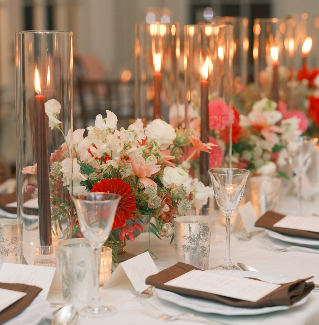 Elegant dining table with smoked-glass candle holders, lush red-pink floral centerpiece, white china, brown napkins, and silver votive holders, creating a classic, high-end ambiance.