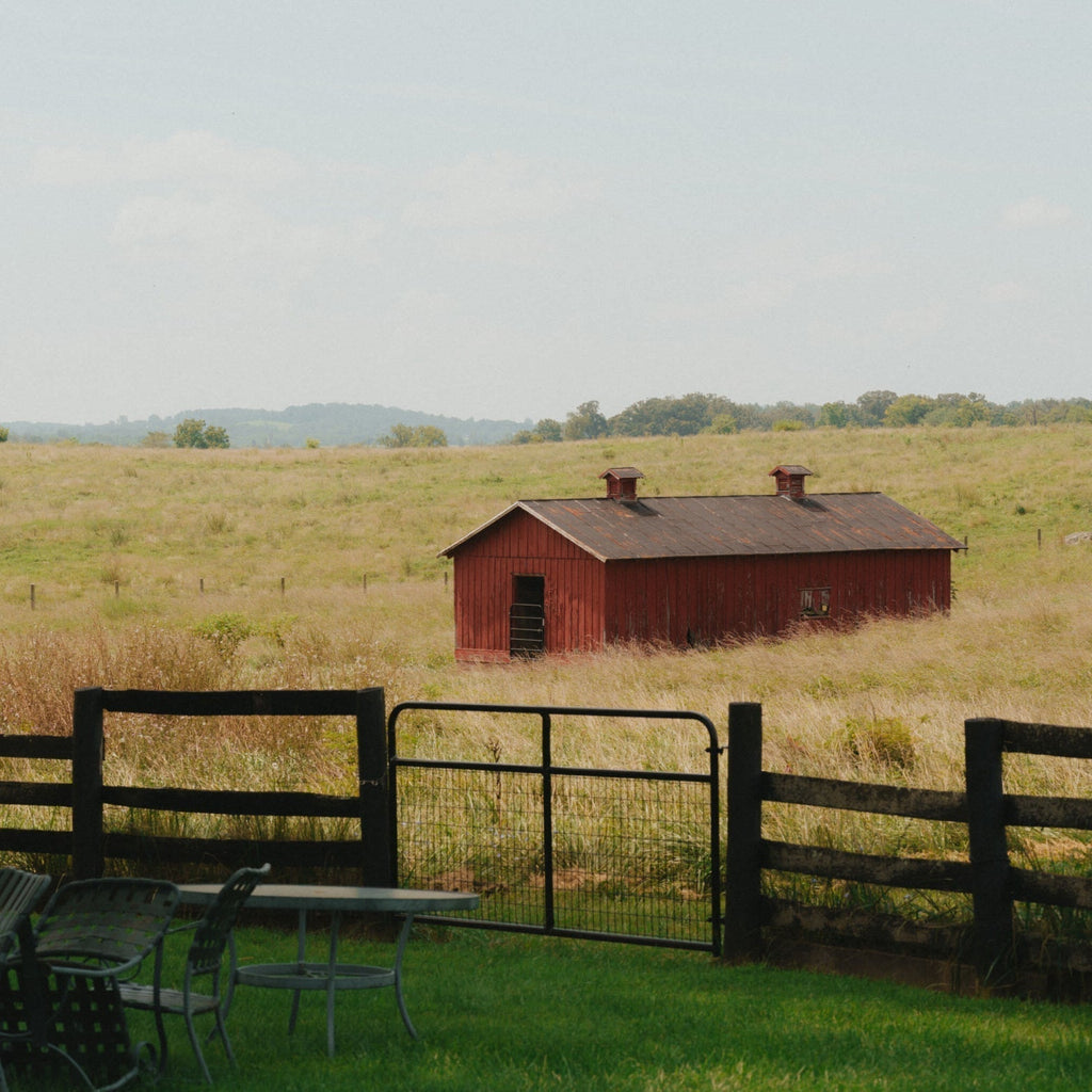 Classic rural scene with an effortless charm: a wooden fence, metal gate, outdoor chairs, table, rustic barn, and rolling hills under a partly cloudy sky.