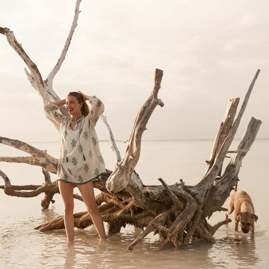 A woman in a classic, effortless tunic with a floral print enjoys a fun beach moment with a dog, embodying high-end, relaxed style.
