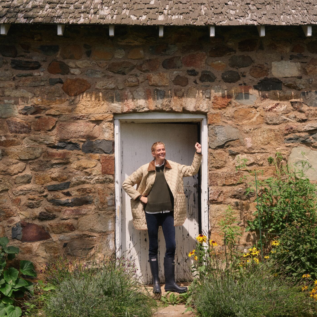 Person in a classic, quilted beige coat, stands in rustic doorway surrounded by flowers, embodying Tuckernuck's effortless, fun, and high-end style.