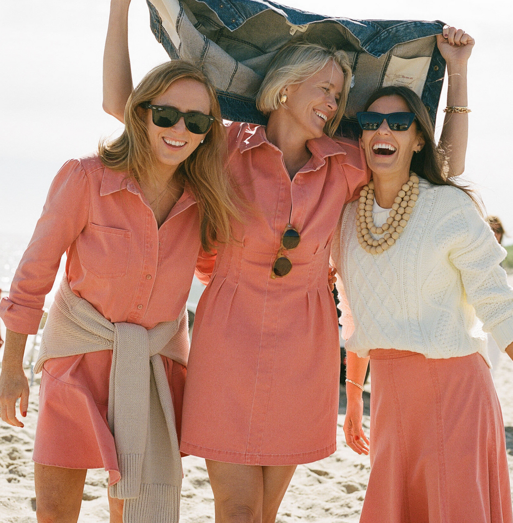 Three women in coral outfits enjoy a sunny beach day, exuding classic, effortless style with fun, high-end fashion from Tuckernuck.