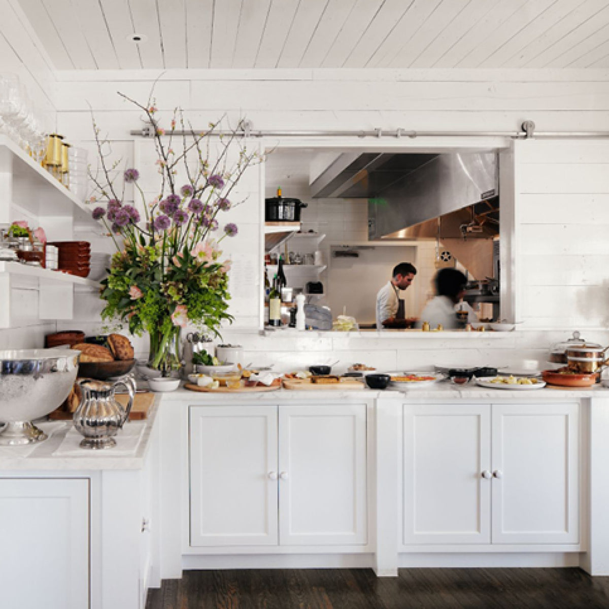 A classic, high-end kitchen pass line featuring an effortless buffet setup with floral arrangements, charcuterie, silverware, and glassware; kitchen staff visible behind.