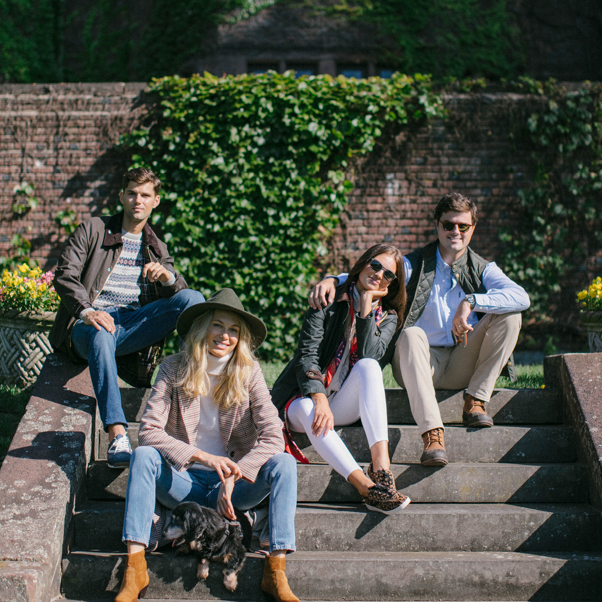 Four adults in classic, effortless outfits sit on garden steps. A woman in a hat pets a dog, embodying high-end, fun Tuckernuck style.