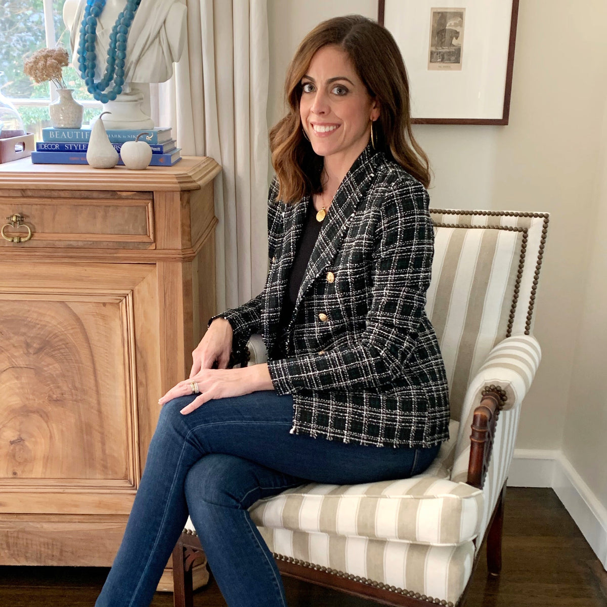 A woman in a classic, high-end tweed blazer, seated in an elegant armchair, embodies effortless style and fun amidst chic decor elements.