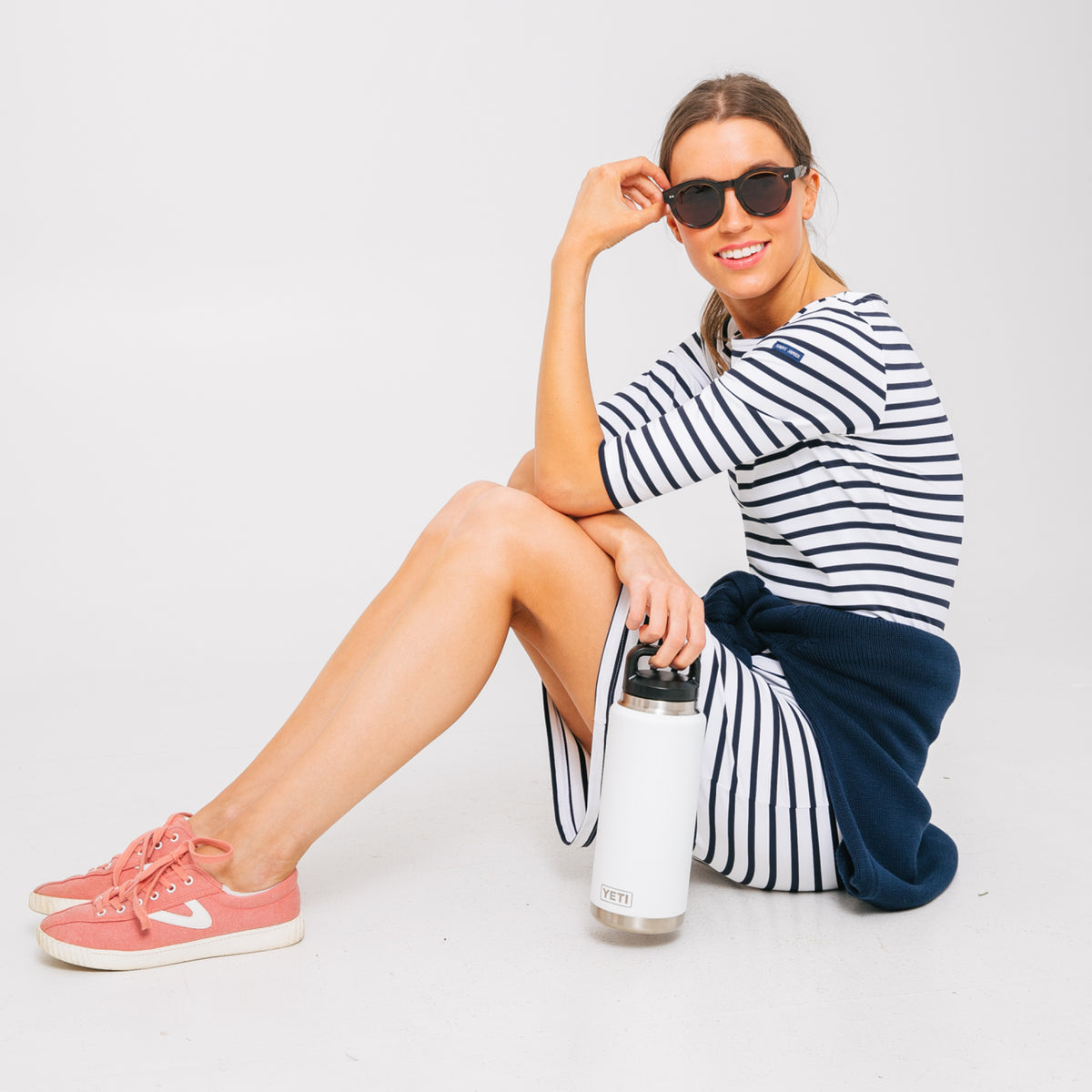 Woman in a classic white striped dress and coral sneakers, effortlessly holding a high-end YETI bottle, exuding fun and sporty-casual style.