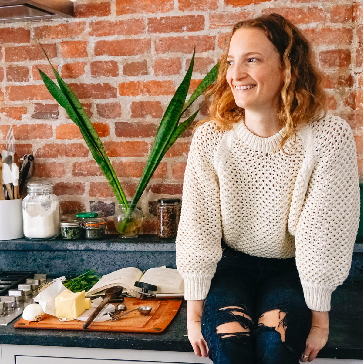 Woman in classic cream sweater and jeans on a kitchen counter with a cutting board, cookbook, and glass jars, embodying effortless, high-end style.