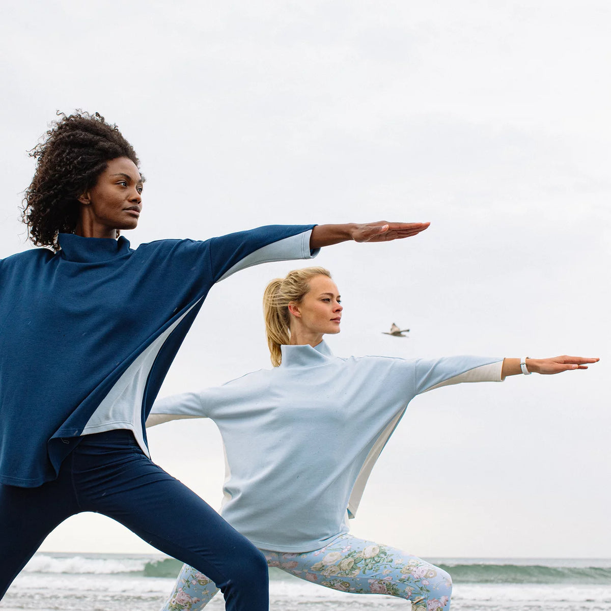 Two women in Warrior II pose on a beach, showcasing classic, effortless yoga wear; one in a blue top, the other in floral leggings.