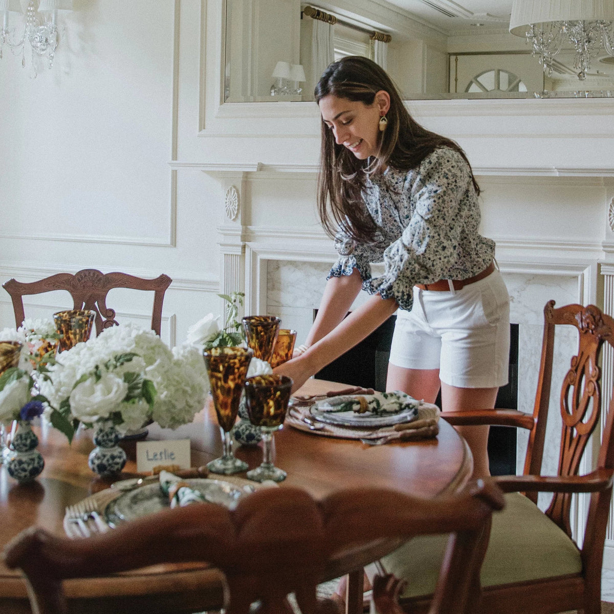 A young woman arranges an effortless, high-end tablescape with classic porcelain vases, amber goblets, and handwritten place cards on a polished wooden dining table.