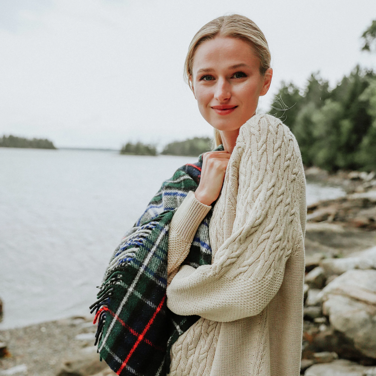Young woman in a classic, cream cable-knit sweater and tartan blanket, enjoying an effortless, high-end moment by a tranquil lakeside.