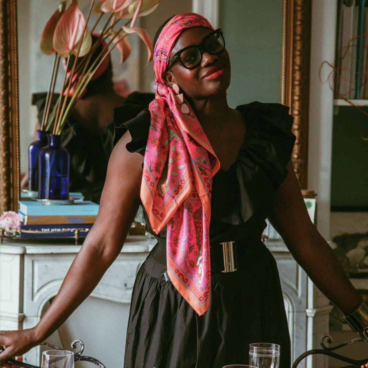 A person in a classic black dress, bold glasses, and a fun scarf stands in an elegant setting with a high-end mantelpiece and table arrangement.