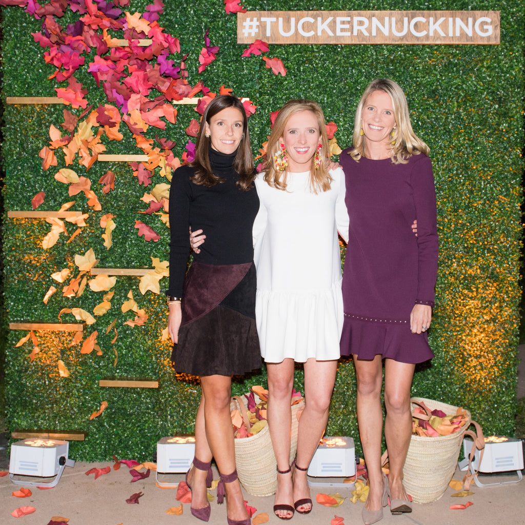 Three women in classic, effortless fall outfits stand before a decorative wall, embodying high-end, fun style with baskets of faux autumn leaves.