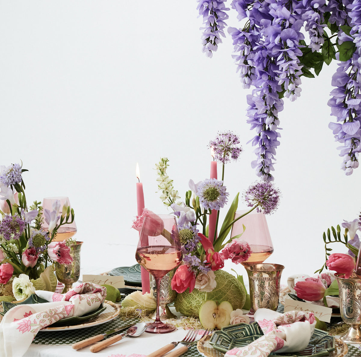 Effortlessly chic spring tablescape with cascading wisteria, pastel blooms, pink taper candles, rose-tinted glassware, and layered plates, creating a classic, high-end garden-party vibe.