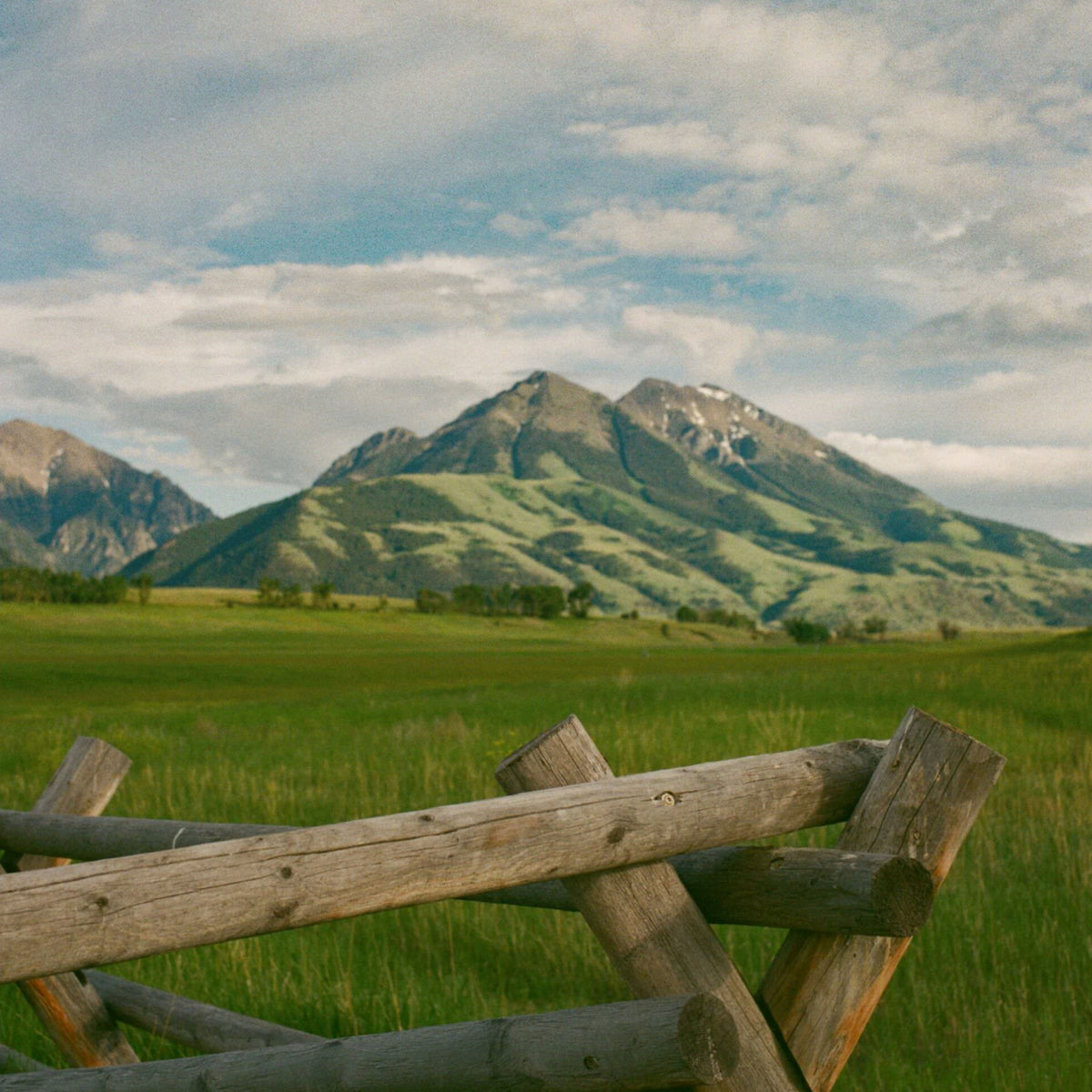 A classic split-rail fence borders a lush meadow, with a mountain backdrop under a partly cloudy sky, embodying effortless beauty and high-end adventure.
