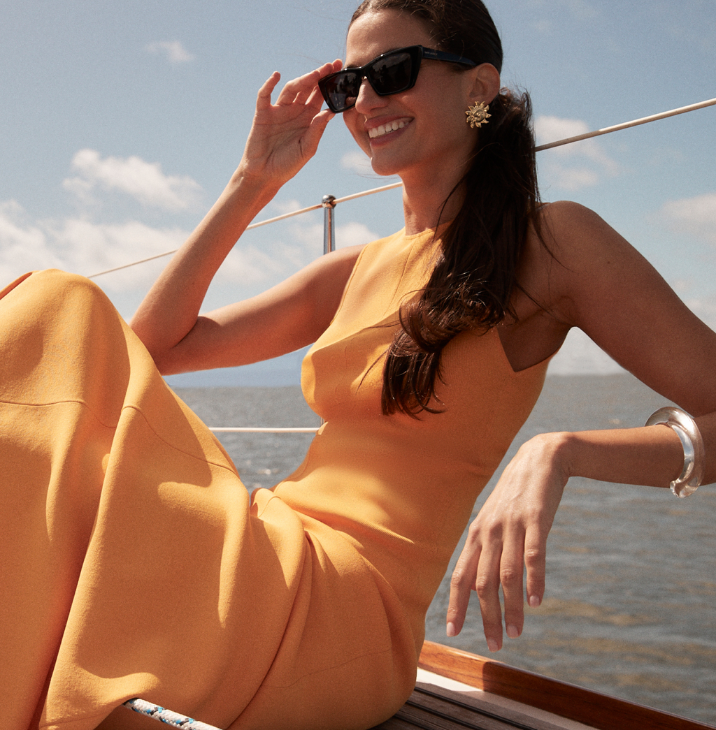 A young woman in a classic golden-yellow dress lounges effortlessly on a sailboat, wearing fun black sunglasses and high-end accessories, exuding confidence.