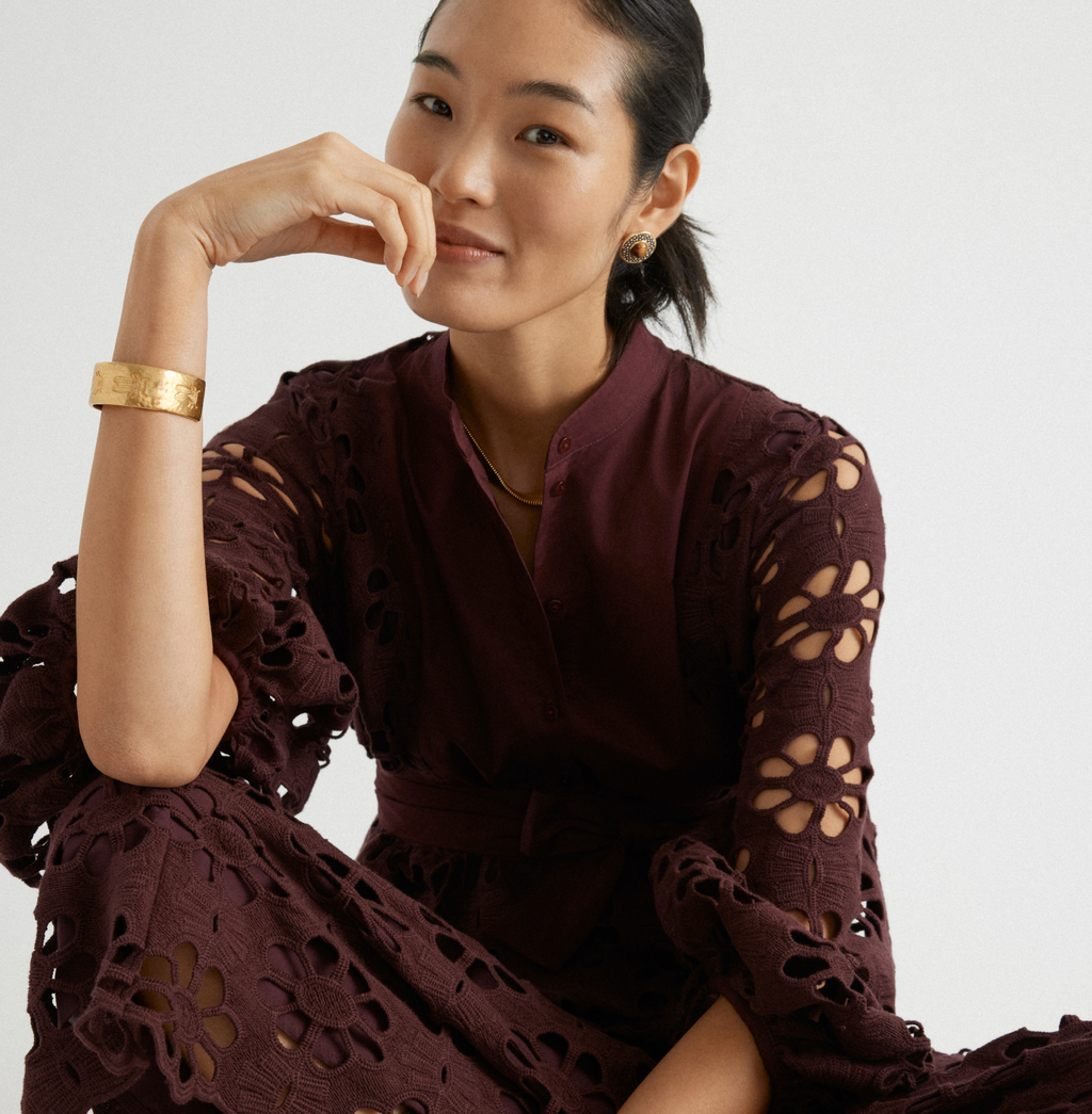 Young woman in a classic, deep burgundy dress with floral cutouts, accessorized with a textured gold cuff, necklace, and earrings, exuding an effortless, high-end elegance.