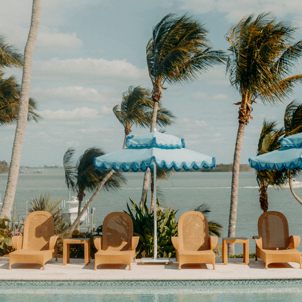 Classic poolside scene with wicker lounge chairs, bright blue umbrellas, and lush palms by a tropical waterway, reflecting effortless, high-end relaxation and fun.