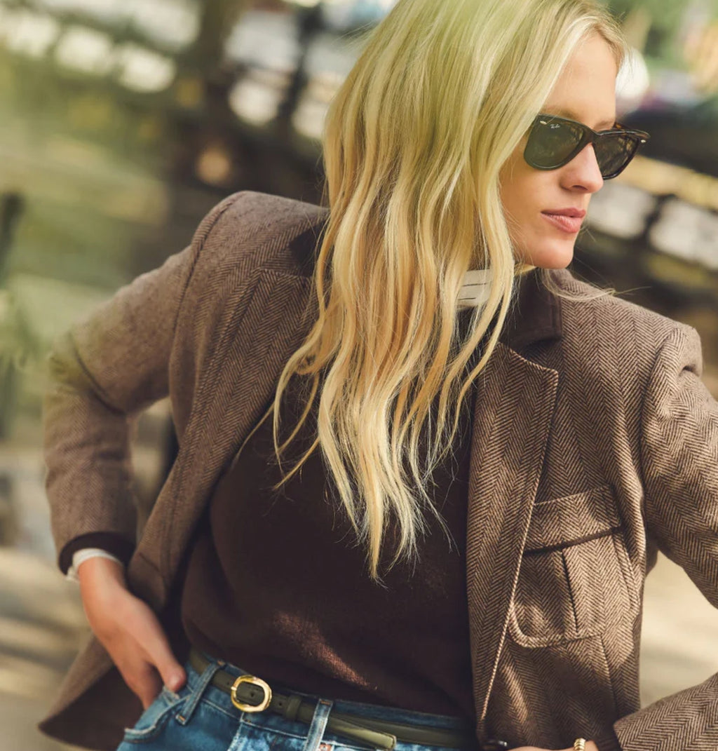 Young woman in classic herringbone blazer and high-waisted jeans, exuding effortless style with sunglasses, embodying Tuckernuck's fun, high-end fashion ethos.