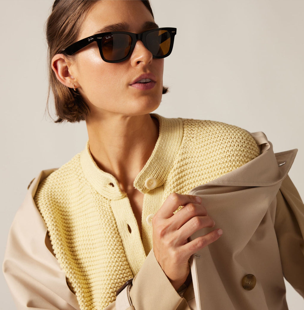 Close-up of a woman wearing classic black sunglasses, an effortless light yellow knit top, and a fun beige trench coat, embodying a high-end look.