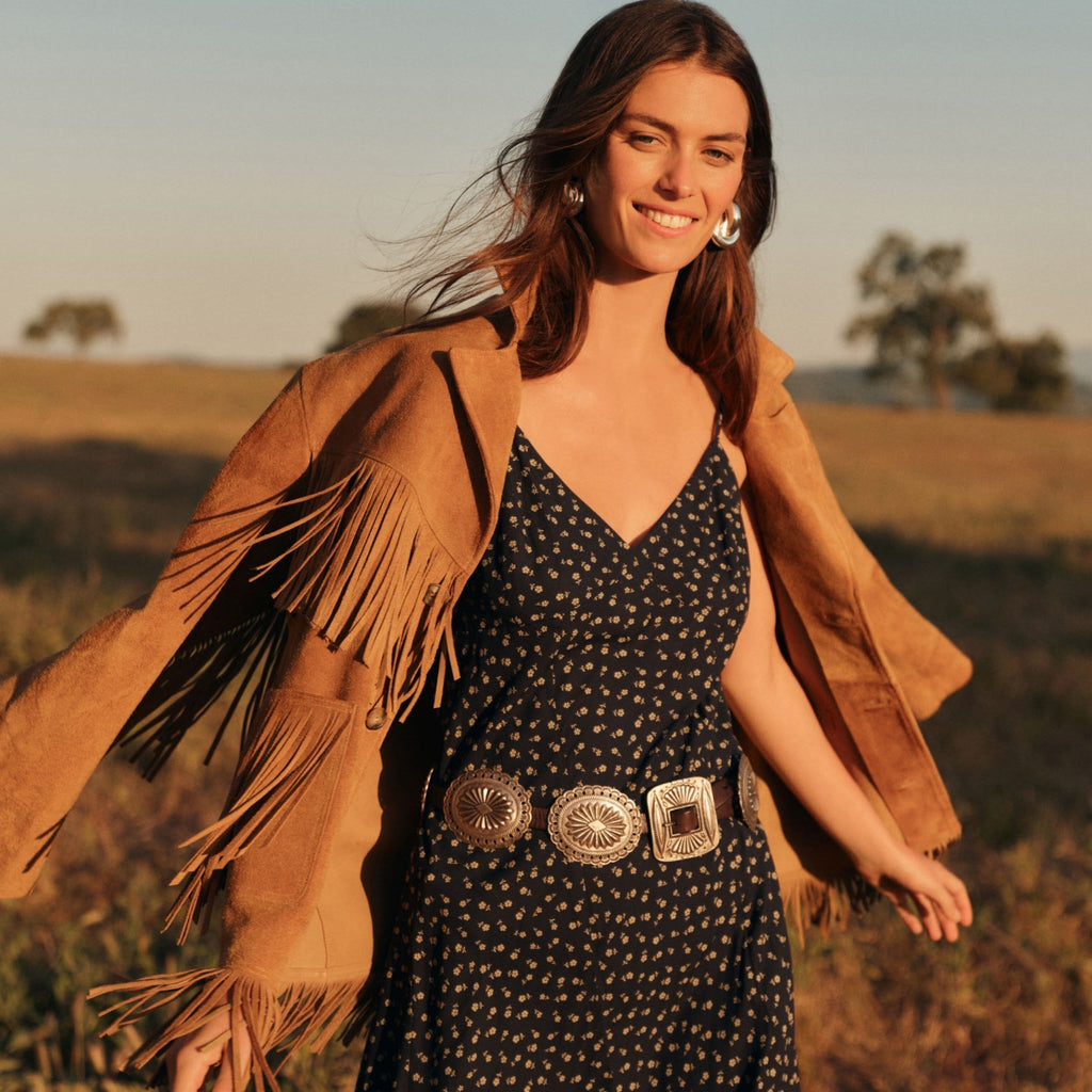 Young woman in a sunlit meadow, wearing an effortlessly cool tan fringe jacket, floral navy dress, and high-end silver accessories, embodying classic, fun style.