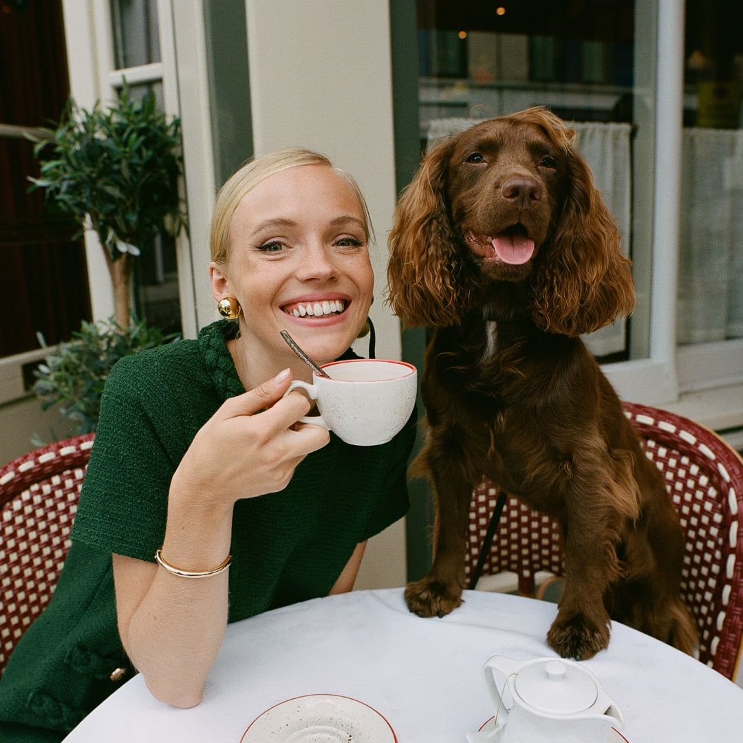A young woman with a classic, effortless style enjoys tea with her playful spaniel at a high-end outdoor café, embodying a fun, confident lifestyle.