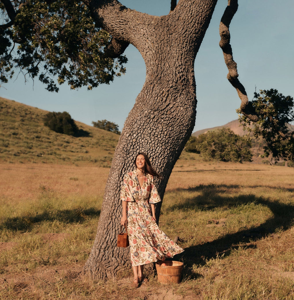 A young woman in a classic floral dress stands effortlessly against a tree, holding a woven basket, embodying Tuckernuck's fun, high-end style.
