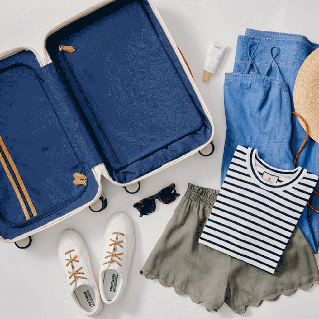Flat-lay packing scene with a classic navy suitcase, white sneakers, tortoiseshell sunglasses, denim dress, straw hat, striped T-shirt, olive shorts, and sunscreen for an effortless, fun trip.