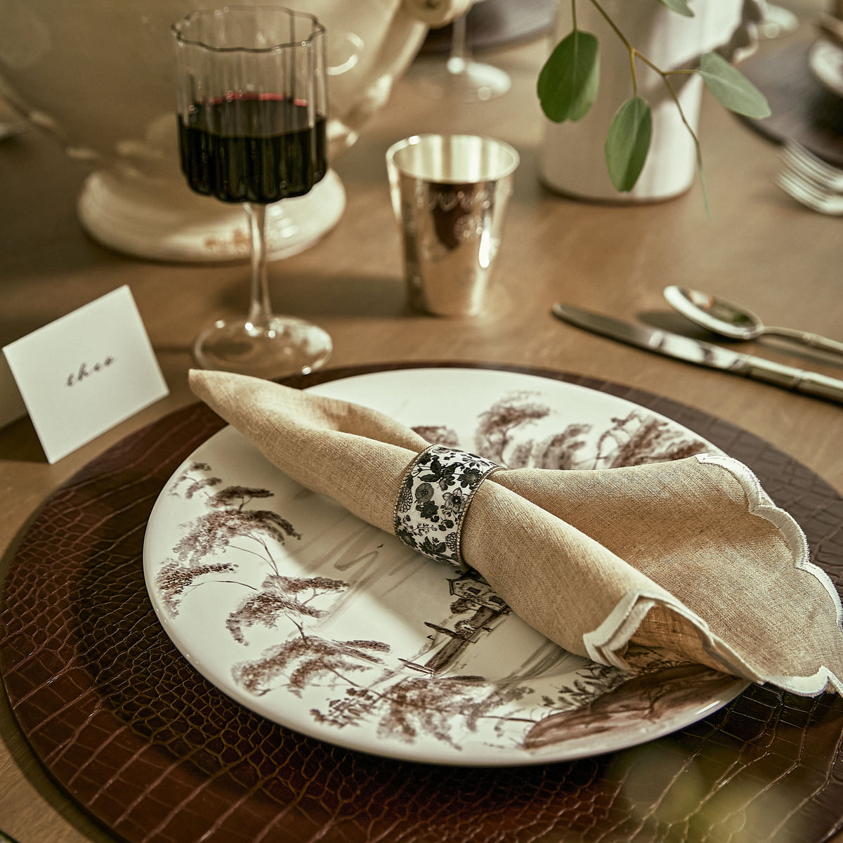 Elegant wooden table setting: classic porcelain plate with sepia pastoral scene, beige napkin, floral ring, red wine glass, cutlery, eucalyptus greenery. Effortless, high-end charm.