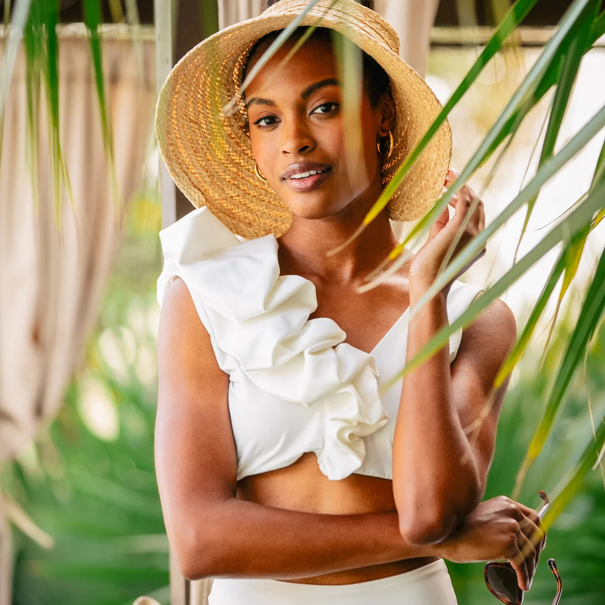 Young woman in a classic, effortless style wearing a wide-brimmed straw hat, gold hoop earrings, and a white one-shoulder ruffled bikini top, exuding high-end resort vibes.