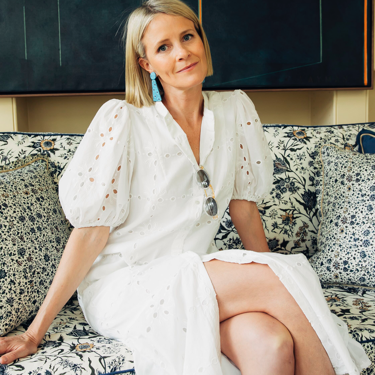 Woman in a classic white eyelet dress with puffed sleeves and turquoise earrings, sitting on a floral sofa, embodying effortless style and high-end sophistication.