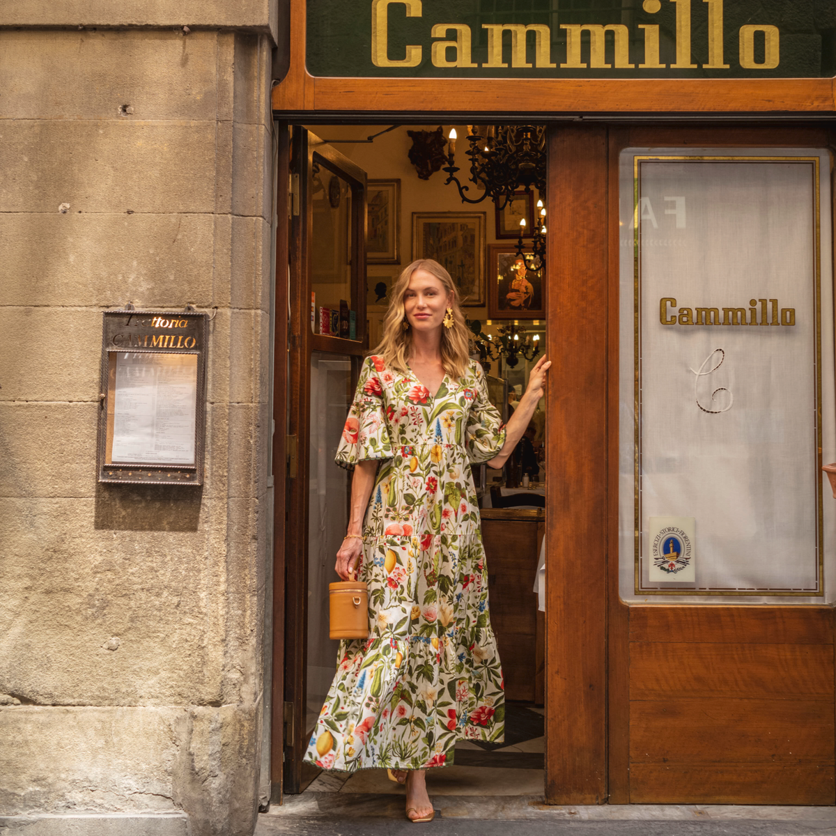 Woman in a classic, floral dress with a tan handbag stands at charming Cammillo café doorway, exuding effortless, high-end style and a fun, inviting atmosphere.