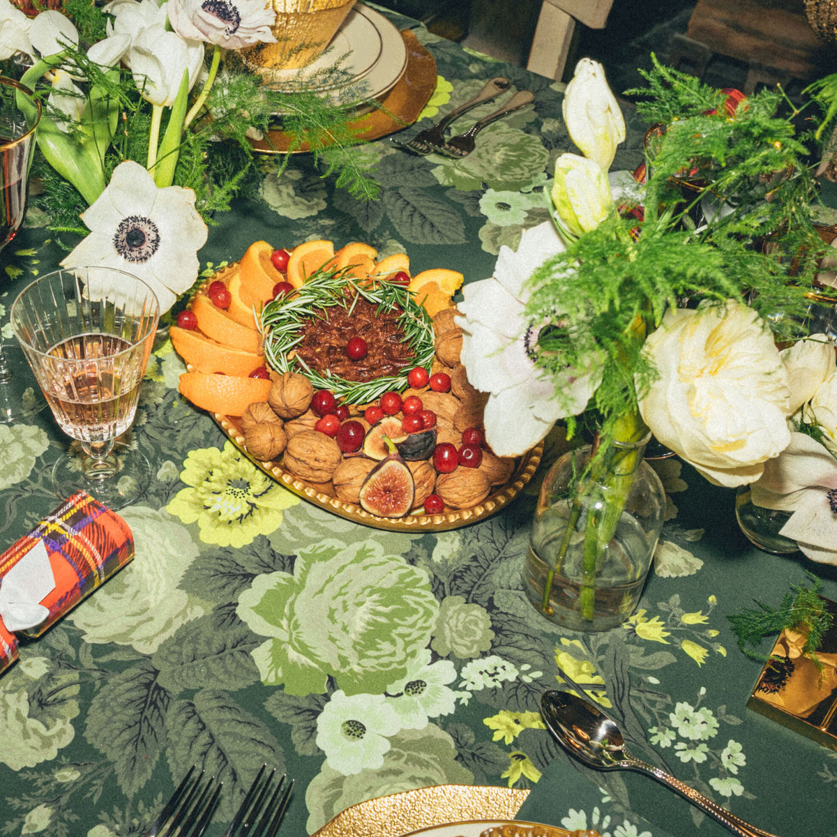 Festive tabletop with classic, high-end appetizers: gold-rimmed platter of walnuts, figs, citrus; crystal wine glass; polished silverware; white flowers; plaid-wrapped favor. Effortless holiday elegance.