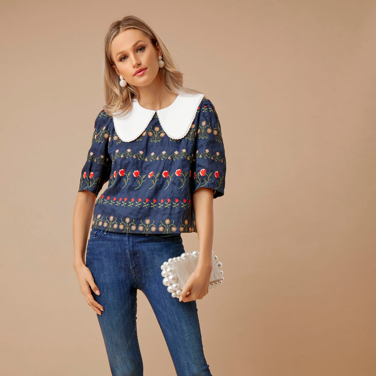 Model in slim-fit jeans and a navy blouse with floral embroidery, classic Peter-Pan collar, holding a pearl-adorned clutch; embodies effortless, high-end style.