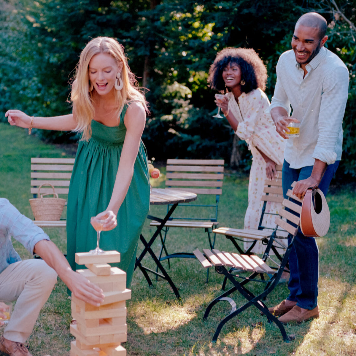 Four friends enjoy a classic, high-end outdoor gathering, playing a giant Jenga game with effortless style and fun, surrounded by a relaxed picnic atmosphere.