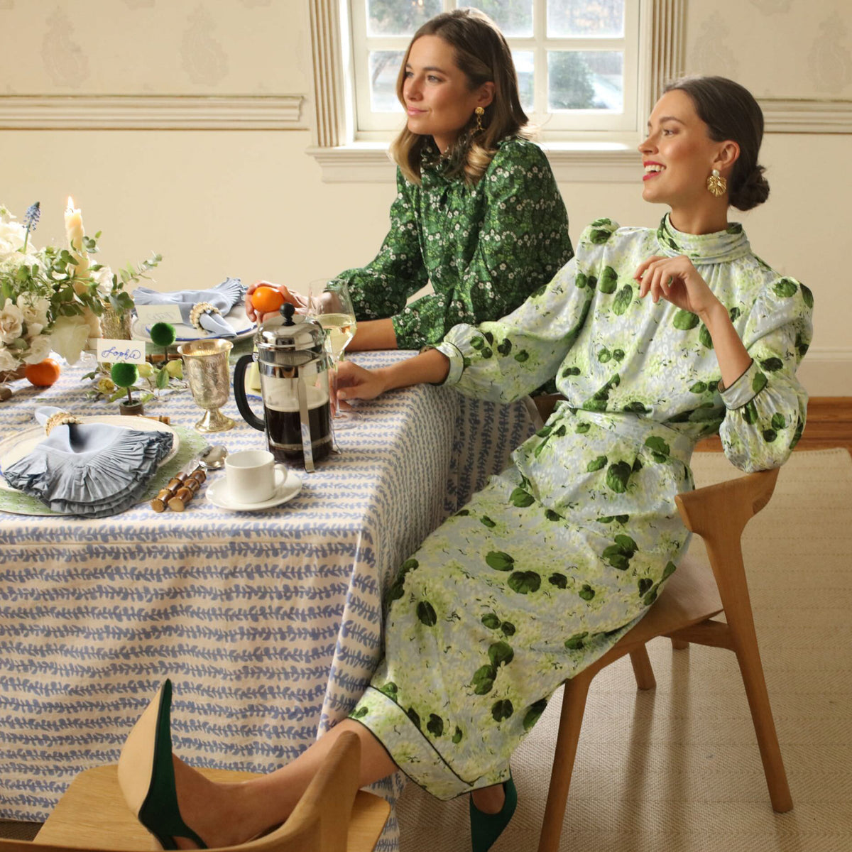 Two women in classic green floral dresses, enjoying an effortless, high-end table setting with coffee, citrus decor, and elegant glassware for a fun gathering.