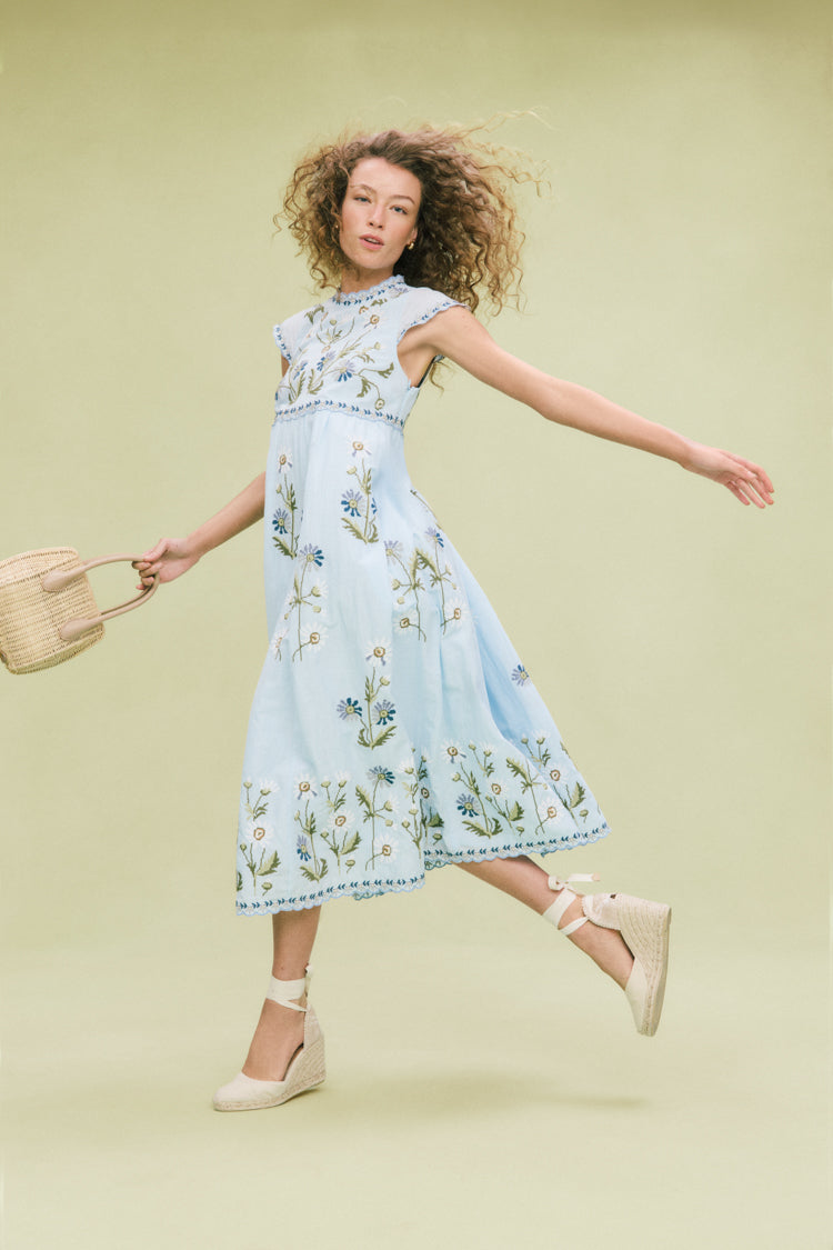 Young woman in motion, showcasing classic Ivory Carina Espadrilles with self-tie ankle straps, effortlessly styled with a floral midi dress and woven straw handbag.
