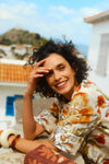 Woman gazing to the side with a picturesque Greek village as her backdrop. Woman is wearing a long floral printed dress with long sleeves and has three bangles on her wrist. 