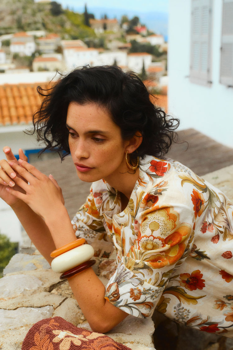 Woman gazing to the side with a picturesque Greek village as her backdrop. Woman is wearing a long floral printed dress. 