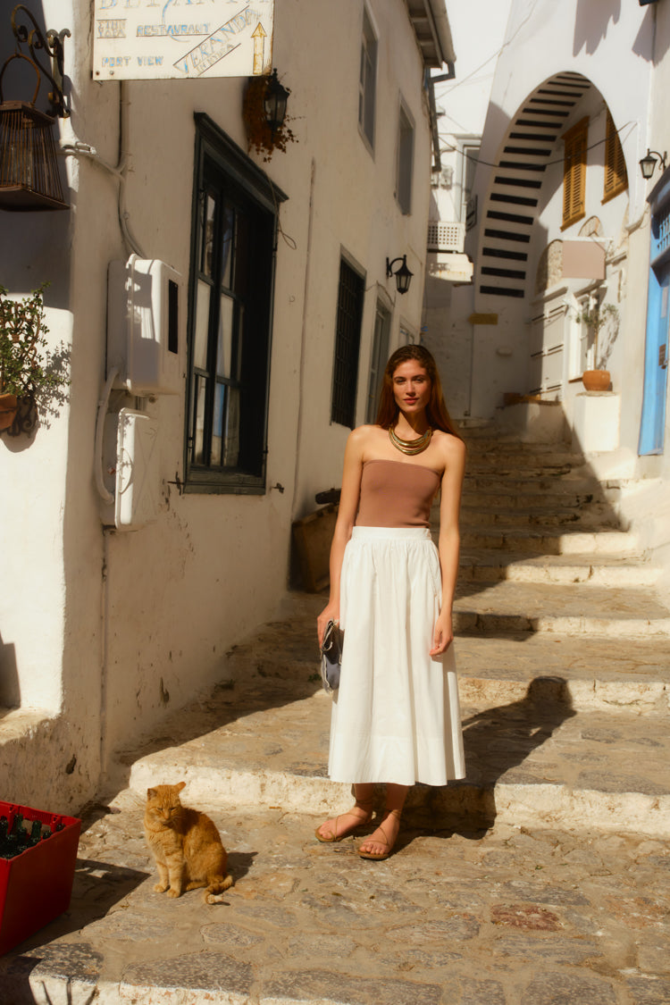 Woman standing on stone stairs in an alleyway wearing a long white skirt and a truffle brown tube top. 