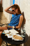 Woman sitting at a cafe table reading a book in a blue and navy crochet dress. 