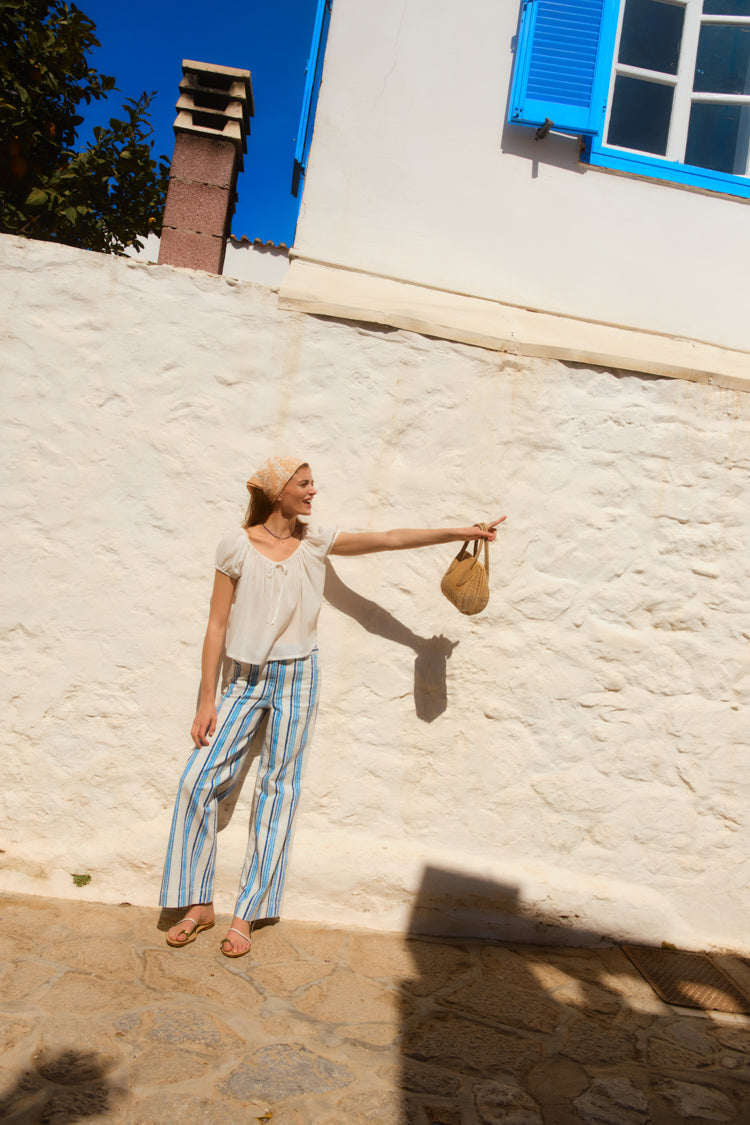 Woman standing in front of a picturesque white wall wearing a white top and blue striped pants. 