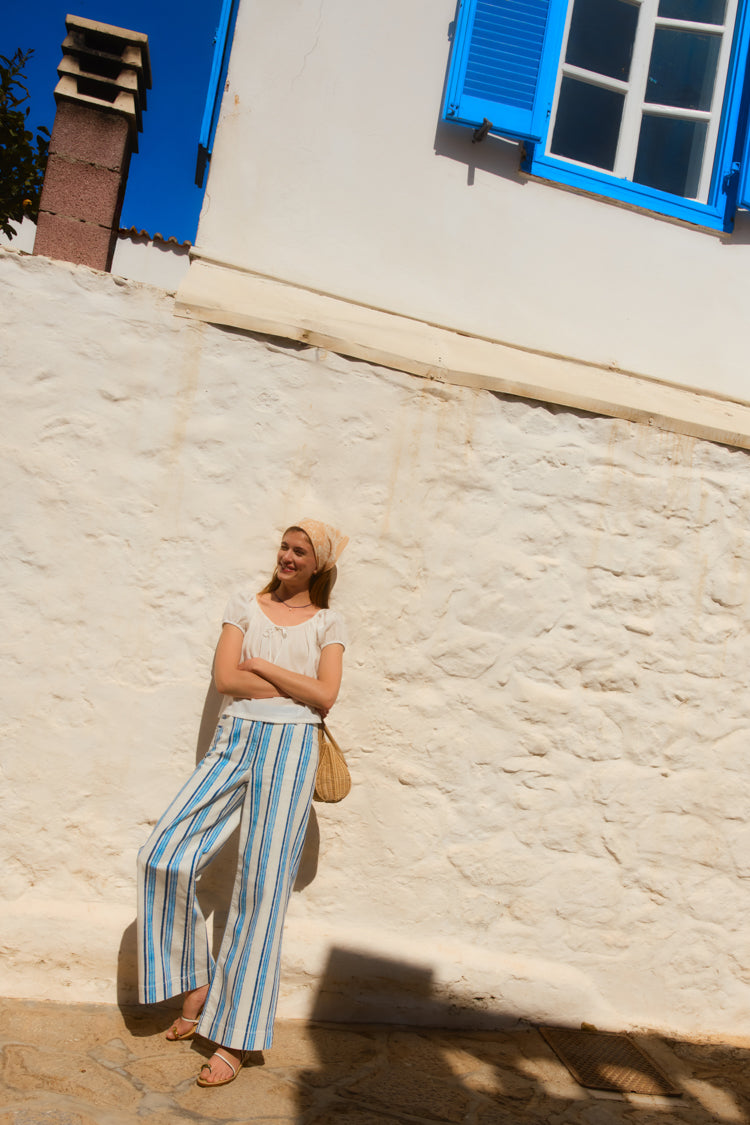 Woman leaning on a picturesque white wall in a white top and blue striped pants. 