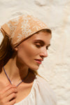 Woman holding a blue necklace with a peach scarf tied around her head. 