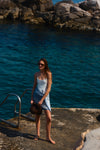 Woman standing by the water in a white one piece swimsuit and a blue pareo tied around her waist. 