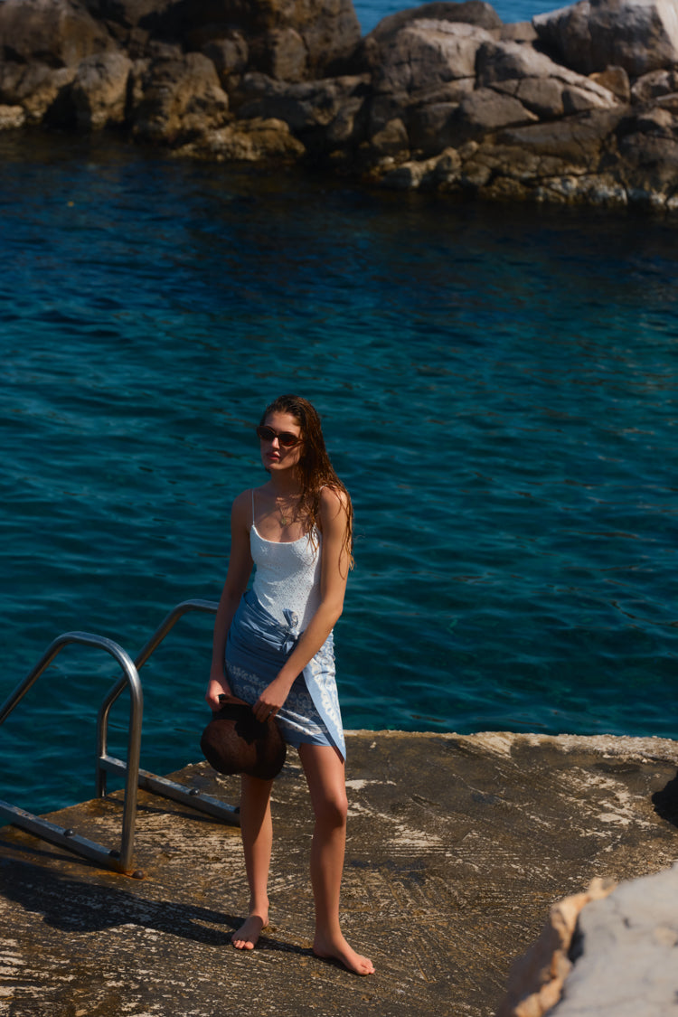 Woman standing by the water in a white one piece swimsuit and a blue pareo tied around her waist. 