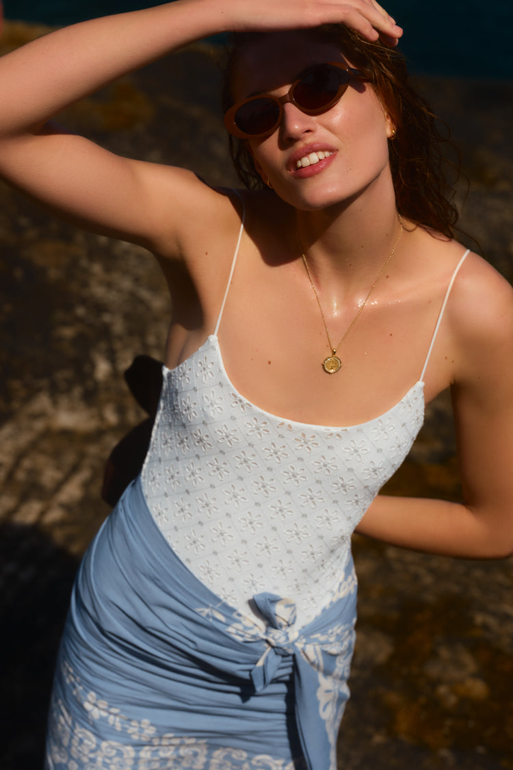 Woman with her hand over her head in a white one piece swimsuit and a blue pareo tied around her waist. 